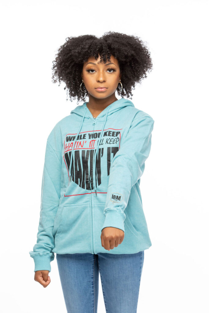 A young woman wearing a blue hoodie, holding one arm in front of her.  On the hoodie is printed, While you keep hatin' it I'll keep makin' it.